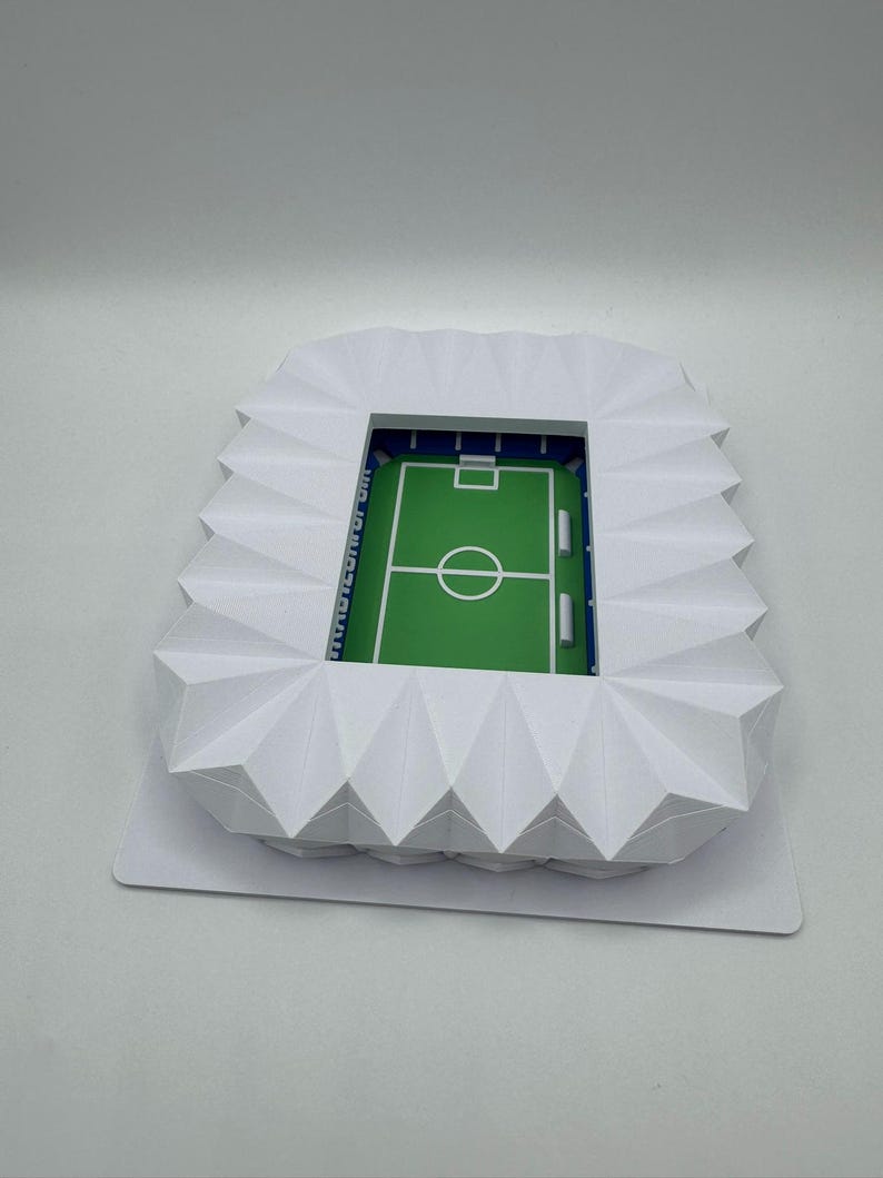 May include: A miniature white paper stadium model with a green field and blue accents. The stadium has a detailed design, including seating areas and a playing field. The text "[UNREADABLE]" is visible on the side of the stadium.