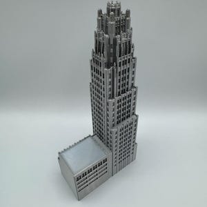 May include: A silver-colored architectural model of a skyscraper with a detailed design. The model features a tall, multi-story tower with a smaller adjacent structure. The building has many windows and intricate details.