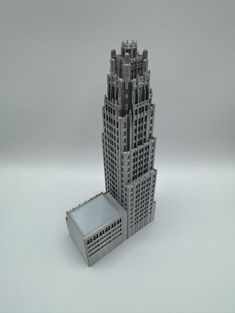 May include: A silver-colored architectural model of a skyscraper with a detailed design. The model features a tall, multi-story tower with a smaller adjacent structure. The building has many windows and intricate details.