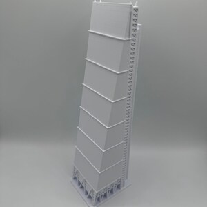 May include: A white architectural model of a tall building with a unique, angled design. The model features multiple rectangular sections stacked on top of each other, with a ladder-like structure on one side. The base has a complex support structure.