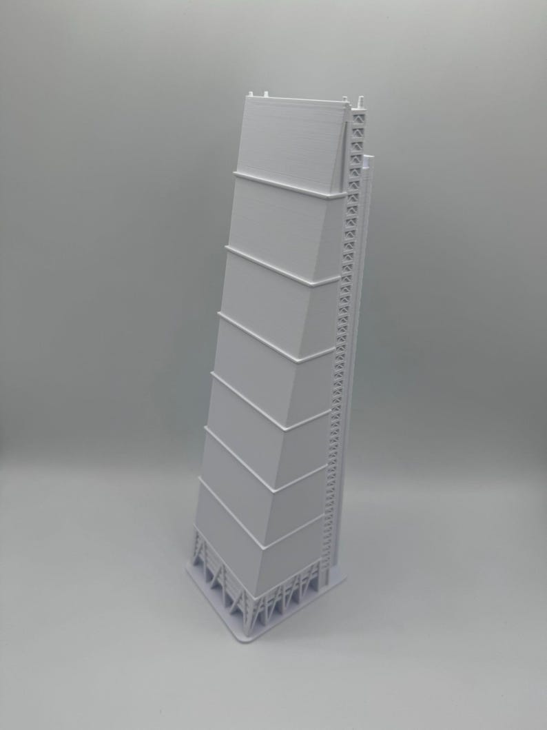 May include: A white architectural model of a tall building with a unique, angled design. The model features multiple rectangular sections stacked on top of each other, with a ladder-like structure on one side. The base has a complex support structure.