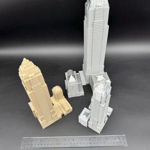 May include: Several miniature skyscraper models in varying shades of gray and tan are arranged on a dark surface. A ruler is placed near the buildings, providing a size reference. The models showcase detailed architectural features.