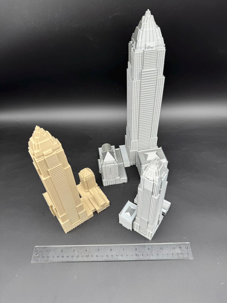 May include: Several miniature skyscraper models in varying shades of gray and tan are arranged on a dark surface. A ruler is placed near the buildings, providing a size reference. The models showcase detailed architectural features.
