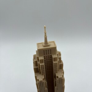 May include: A beige miniature model of a skyscraper with a pointed top. The model is made of plastic and has a detailed facade.