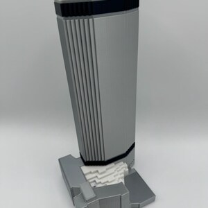 May include: A 3D-printed model of a modern skyscraper. The building is primarily gray with black and white accents. The design features vertical lines and a rectangular base. A small white square is on top.