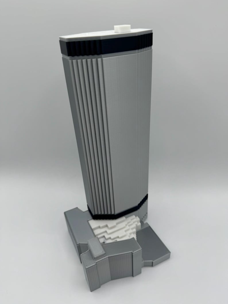 May include: A 3D-printed model of a modern skyscraper. The building is primarily gray with black and white accents. The design features vertical lines and a rectangular base. A small white square is on top.