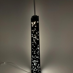 May include: A black, rectangular pendant light fixture with numerous small, bright white lights, resembling a cityscape at night. The light hangs from a white cord and is mounted on a light gray wall.