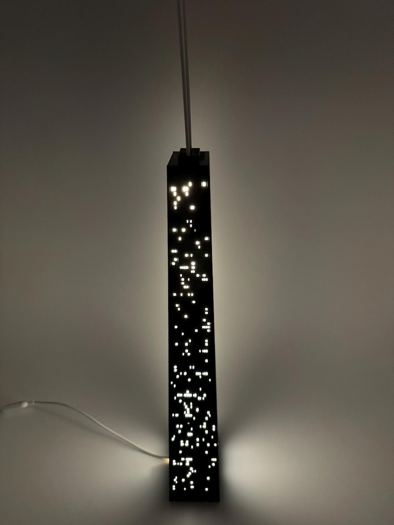 May include: A black, rectangular pendant light fixture with numerous small, bright white lights, resembling a cityscape at night. The light hangs from a white cord and is mounted on a light gray wall.