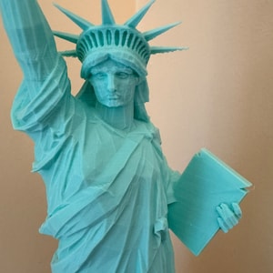 May include: A turquoise blue 3D printed statue of the Statue of Liberty. The statue is holding a tablet in her right hand and her left hand is raised.