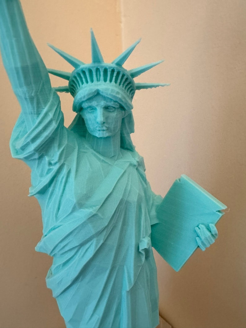 May include: A turquoise blue 3D printed statue of the Statue of Liberty. The statue is holding a tablet in her right hand and her left hand is raised.
