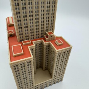 May include: A beige and red architectural model of a skyscraper. The model features multiple rectangular sections with numerous small, dark windows. The roof is a vibrant red, and the overall design is highly detailed.