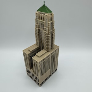 May include: A detailed model of a skyscraper with a beige facade and a green roof. The building has many windows and architectural details, with a small statue on top. The model is set against a white background.