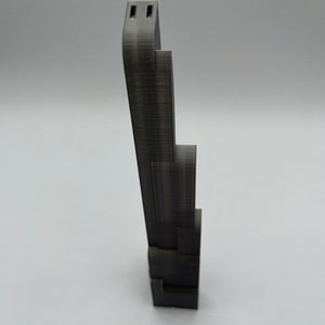 May include: A dark gray, 3D-printed phone stand. The stand has a stepped design with a tall, curved top section featuring two rectangular cutouts. The base is a series of stacked blocks. The stand is designed to hold a phone.