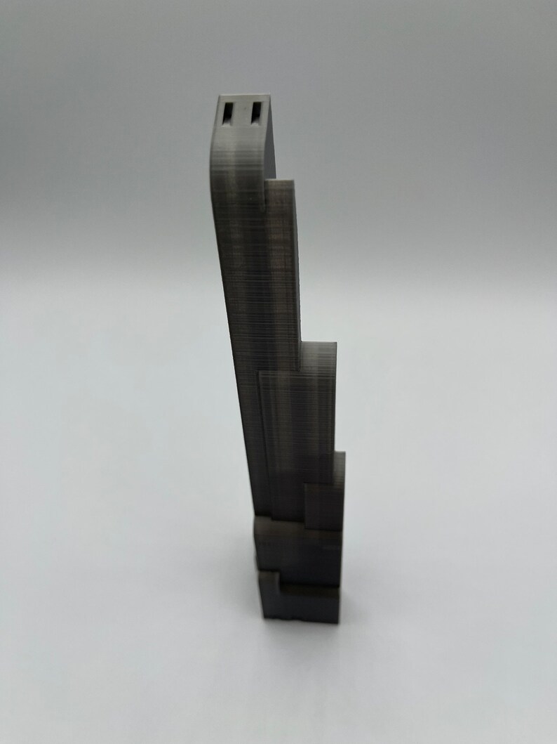 May include: A dark gray, 3D-printed phone stand. The stand has a stepped design with a tall, curved top section featuring two rectangular cutouts. The base is a series of stacked blocks. The stand is designed to hold a phone.