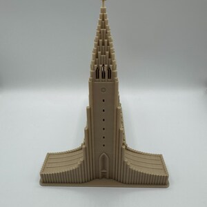 May include: A beige architectural model of a church with a tall, pointed steeple topped with a cross. The building has a series of vertical lines and a rectangular base. The model is made of a solid material.