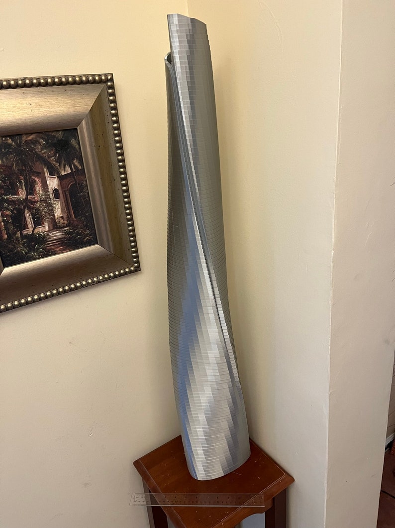 Op de afbeelding: Een zilverkleurig, 3D-geprint model van een hoog, draaiend gebouw. Het model staat op een houten tafel.