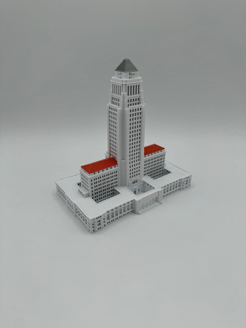 May include: A white 3D printed model of the Los Angeles City Hall building. The building has a tall central tower with a gray roof and red roofs on the wings.