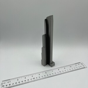 May include: A gray, architectural-style sculpture with a ruler in the foreground. The sculpture has a layered design, resembling a building or structure. The ruler shows measurements in inches, providing scale. The background is plain white.