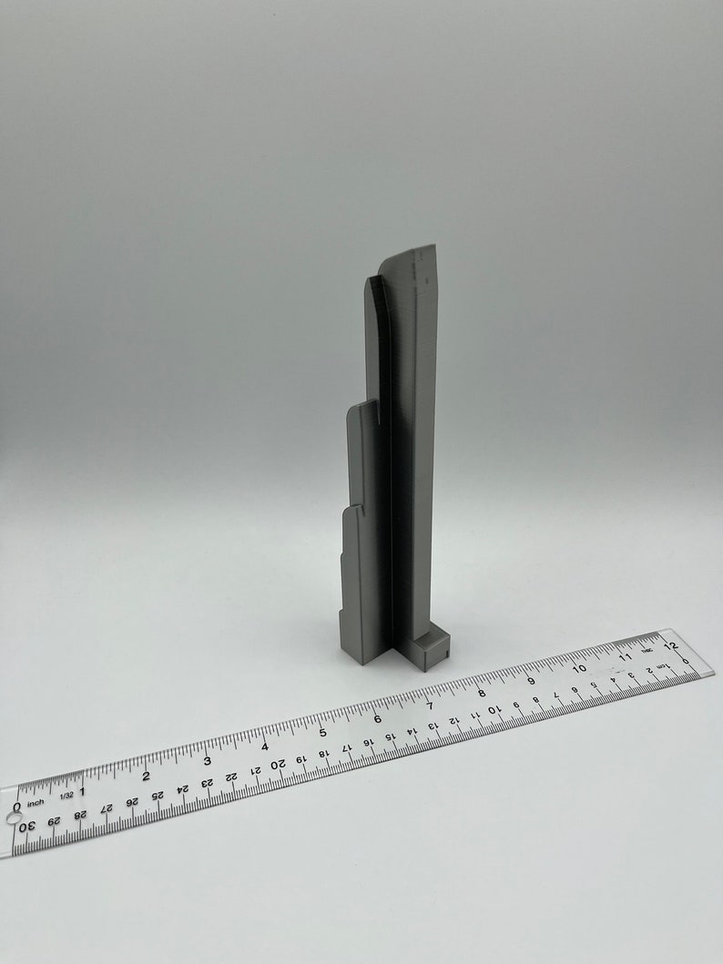 May include: A gray, architectural-style sculpture with a ruler in the foreground. The sculpture has a layered design, resembling a building or structure. The ruler shows measurements in inches, providing scale. The background is plain white.