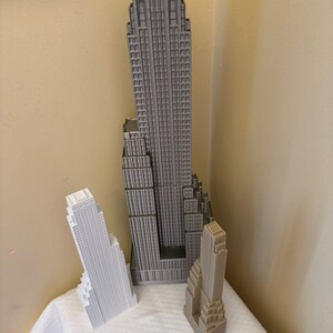 May include: Three miniature skyscraper models in varying shades of gray and white are displayed on a white surface. A ruler is placed in front of the models. The tallest model is approximately 12 inches tall.