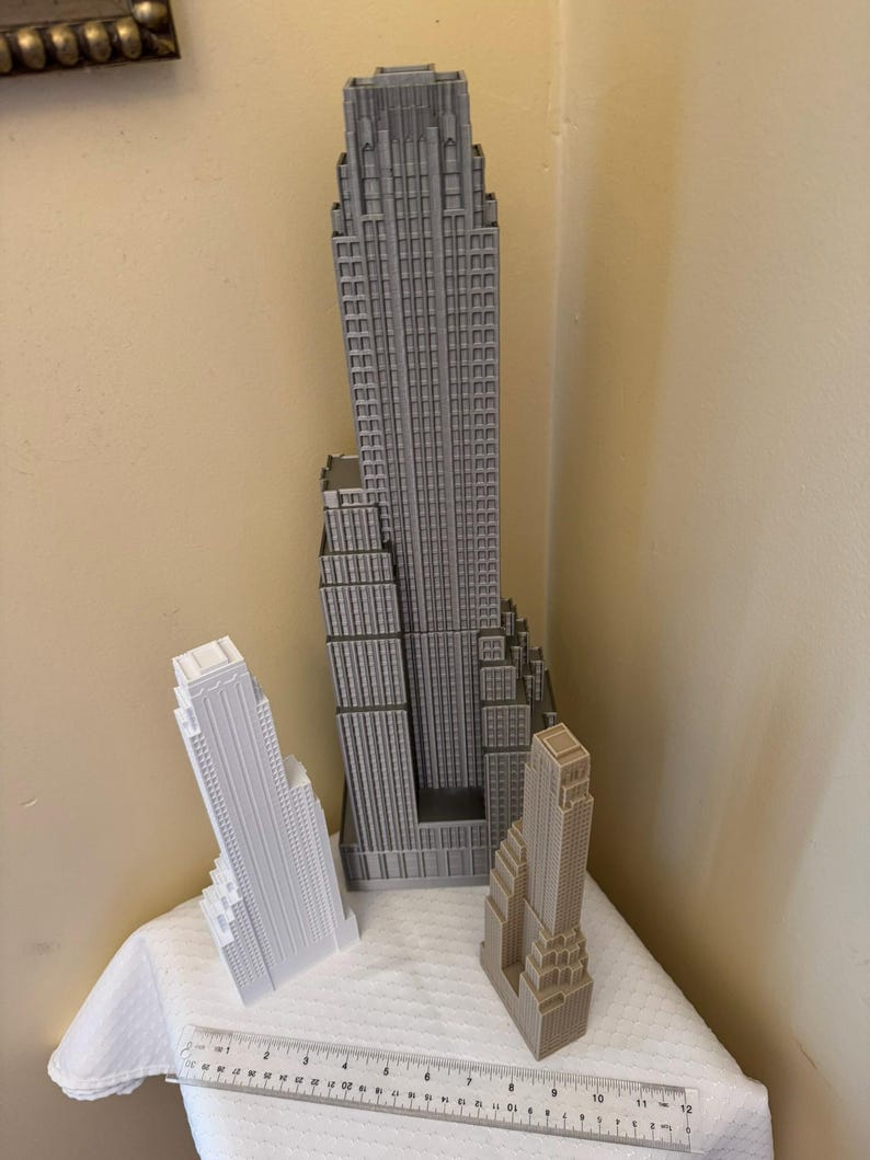 May include: Three miniature skyscraper models in varying shades of gray and white are displayed on a white surface. A ruler is placed in front of the models. The tallest model is approximately 12 inches tall.