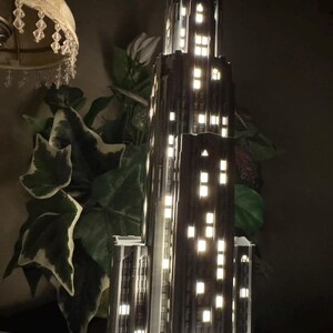 May include: A dark gray, multi-tiered skyscraper model lamp featuring a vertical architectural design with numerous rectangular cutouts that emit a warm, bright white light, simulating illuminated windows in a tall city building at night.