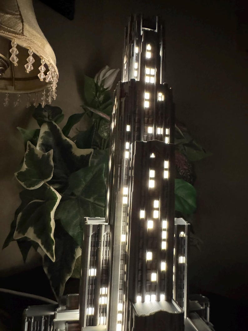 May include: A dark gray, multi-tiered skyscraper model lamp featuring a vertical architectural design with numerous rectangular cutouts that emit a warm, bright white light, simulating illuminated windows in a tall city building at night.