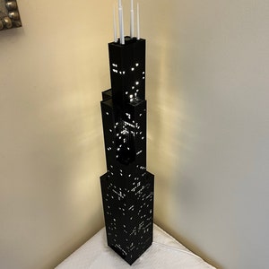 May include: A black LEGO model of the Willis Tower, also known as the Sears Tower, with white antenna details. The model is lit up from within, creating a glowing effect.