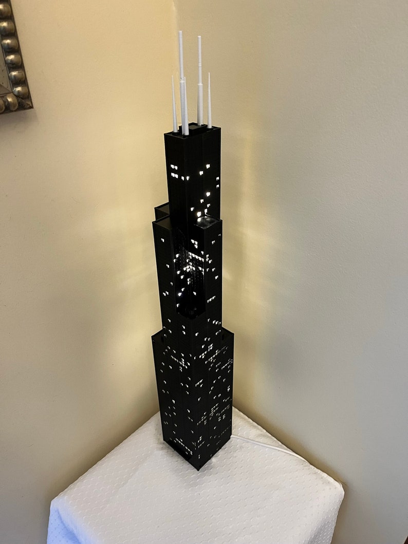 May include: A black LEGO model of the Willis Tower, also known as the Sears Tower, with white antenna details. The model is lit up from within, creating a glowing effect.