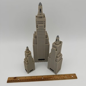 May include: Three beige miniature skyscraper models of varying sizes, with detailed architectural features. A wooden ruler, approximately 12 inches long, is placed in front of the buildings. The buildings have a detailed facade and a pointed top.