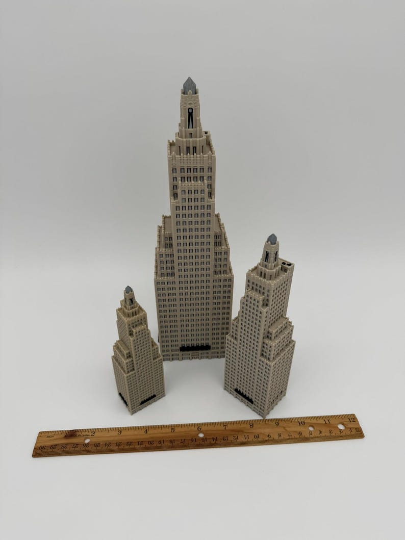 May include: Three beige miniature skyscraper models of varying sizes, with detailed architectural features. A wooden ruler, approximately 12 inches long, is placed in front of the buildings. The buildings have a detailed facade and a pointed top.