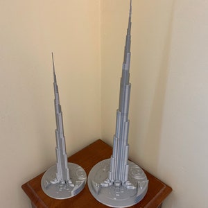 May include: Two silver 3D printed models of the Burj Khalifa, one larger than the other, on a wooden table.
