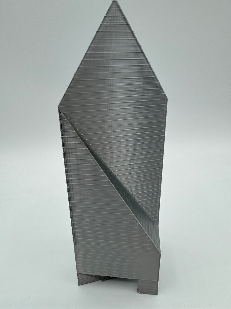 May include: A gray 3D printed model of a tall, slender building with a pointed roof and a geometric design.