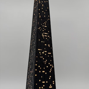 May include: A black skyscraper-shaped lamp with illuminated windows. The lamp has a geometric design and a power cord. The windows emit a warm, yellow light, creating a cozy ambiance. The lamp is a decorative item.