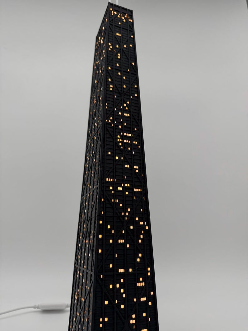 May include: A black skyscraper-shaped lamp with illuminated windows. The lamp has a geometric design and a power cord. The windows emit a warm, yellow light, creating a cozy ambiance. The lamp is a decorative item.