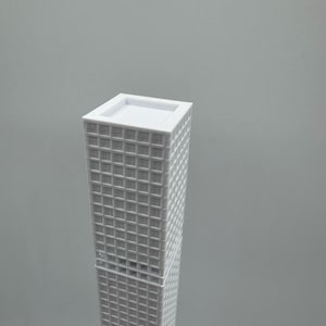 May include: A white miniature skyscraper model with a rectangular base and a flat roof. The building's facade features a grid-like pattern of windows. The model is set against a plain grey background.