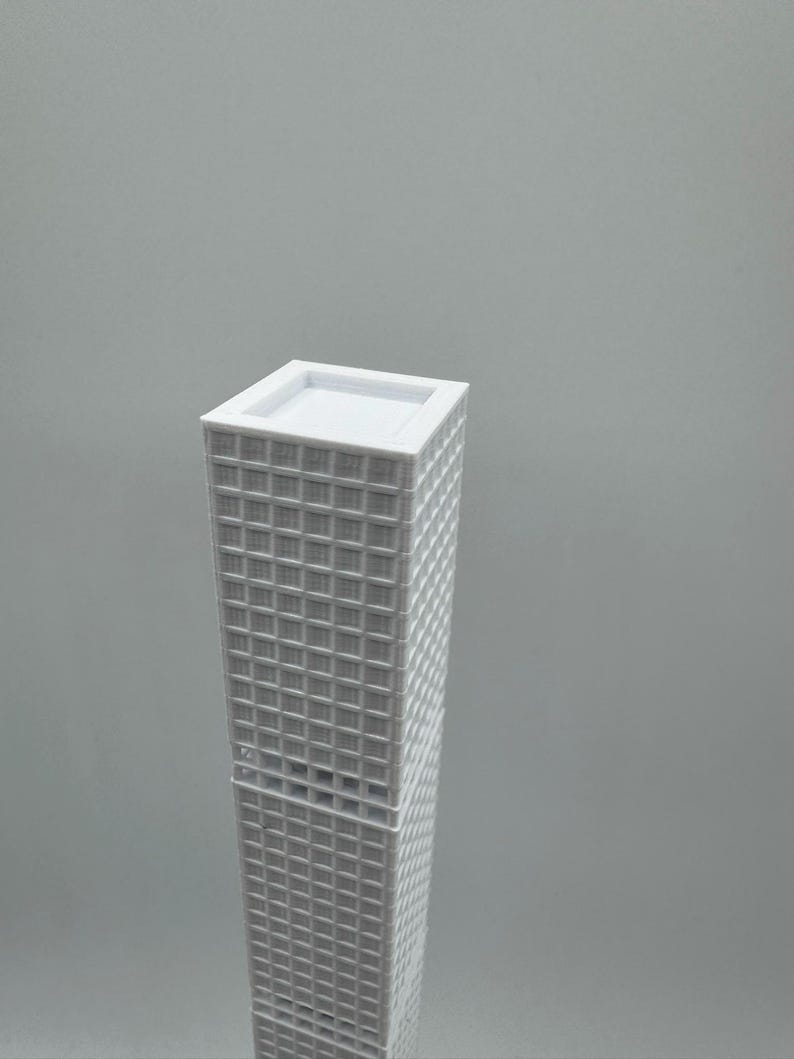 May include: A white miniature skyscraper model with a rectangular base and a flat roof. The building's facade features a grid-like pattern of windows. The model is set against a plain grey background.