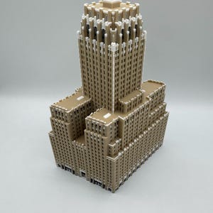 May include: A tan-coloured miniature model of a skyscraper. The building has a tiered design with multiple rectangular sections and a central tower. White accents highlight architectural details, creating a contrast against the tan facade. The model is set against a plain white background.