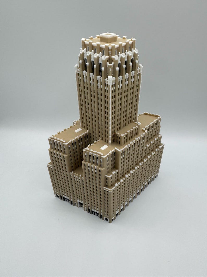 May include: A tan-coloured miniature model of a skyscraper. The building has a tiered design with multiple rectangular sections and a central tower. White accents highlight architectural details, creating a contrast against the tan facade. The model is set against a plain white background.