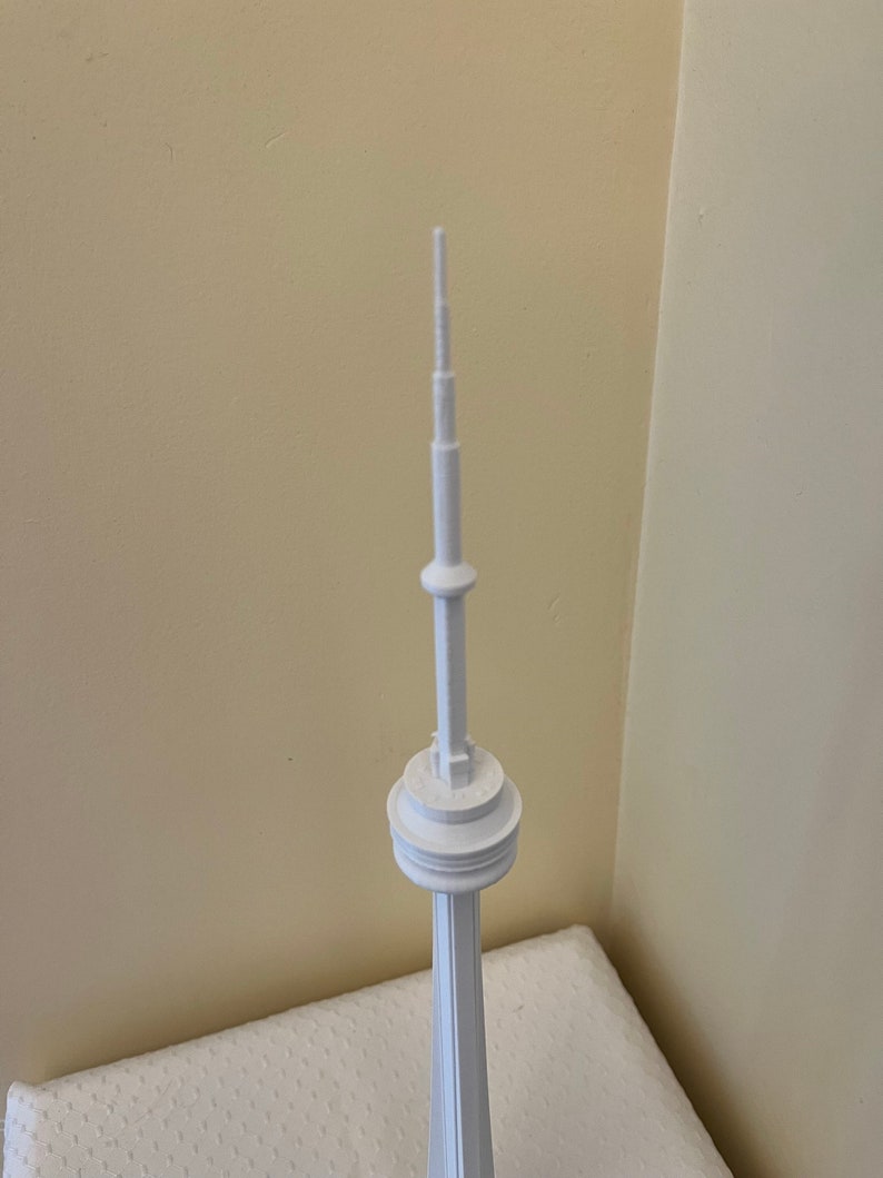 May include: A white miniature replica of the CN Tower. The model features a detailed design, including the tower's distinctive antenna and observation decks. The tower stands tall against a neutral background, showcasing its architectural features.