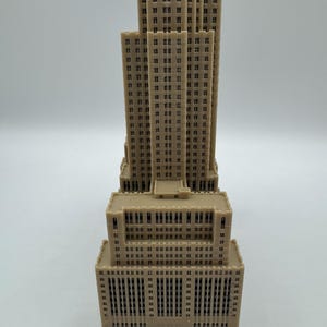 Może przedstawiać: Beżowy miniaturowy model wieżowca z wieloma poziomami i prostokątnymi oknami. Model ma szczeg&oacute;łową fasadę, prezentującą elementy architektoniczne. Projekt budynku obejmuje wysoką centralną wieżę i szerszą podstawę.