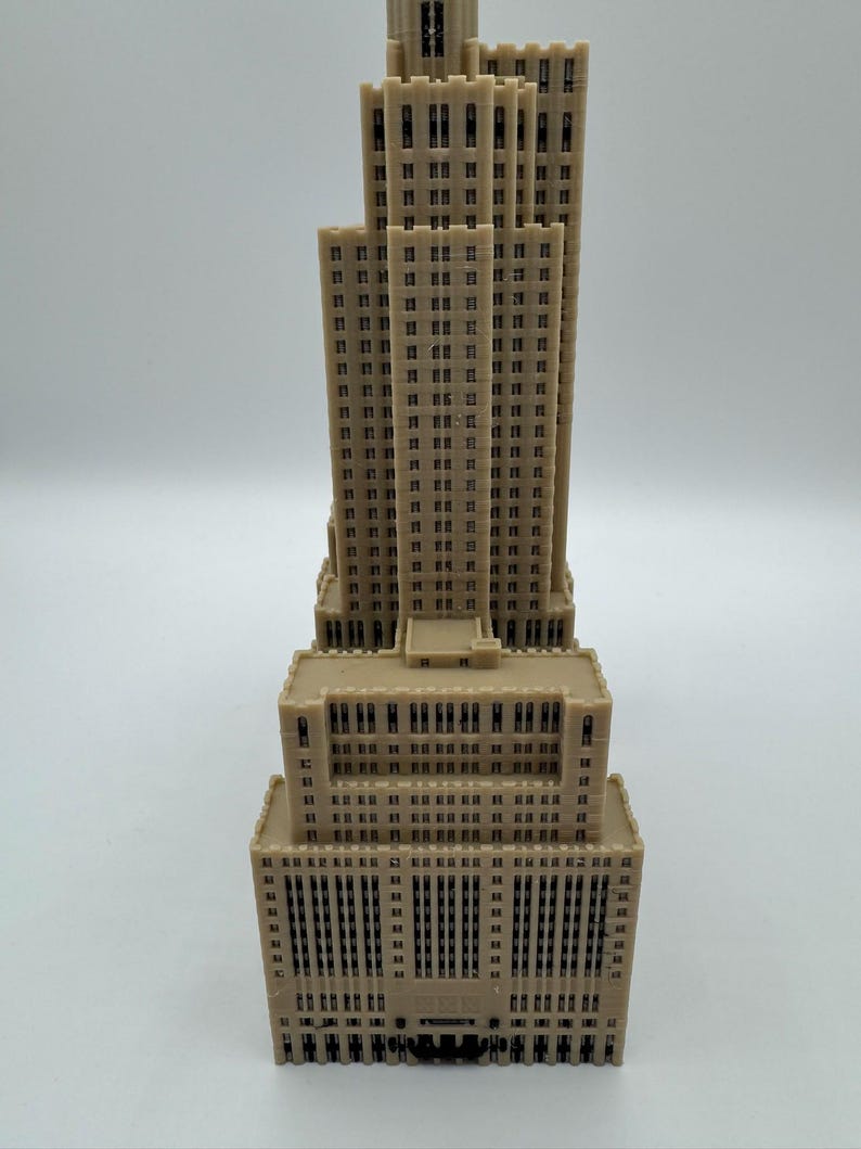 Może przedstawiać: Beżowy miniaturowy model wieżowca z wieloma poziomami i prostokątnymi oknami. Model ma szczeg&oacute;łową fasadę, prezentującą elementy architektoniczne. Projekt budynku obejmuje wysoką centralną wieżę i szerszą podstawę.