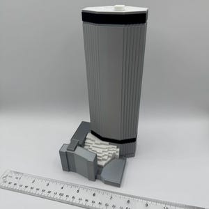 May include: A gray 3D-printed model of a tall building with a white top and black band. The model has vertical lines and sits on a base. A ruler is placed next to the model, showing measurements in inches.