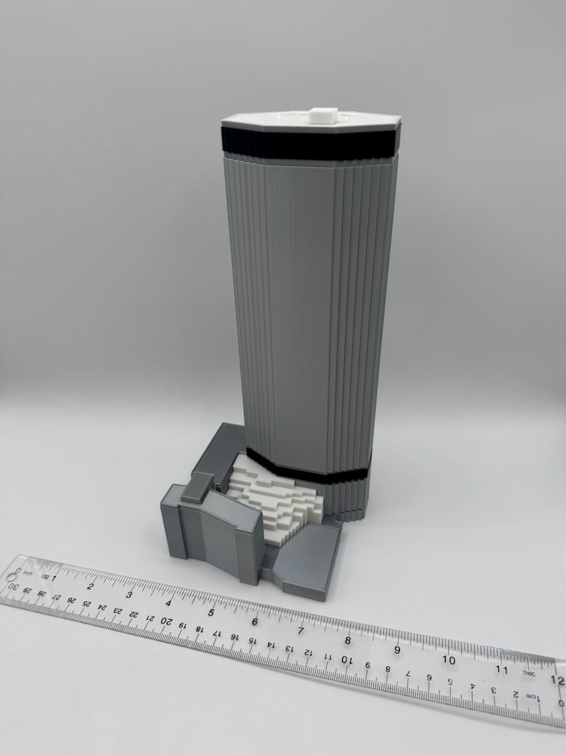 May include: A gray 3D-printed model of a tall building with a white top and black band. The model has vertical lines and sits on a base. A ruler is placed next to the model, showing measurements in inches.