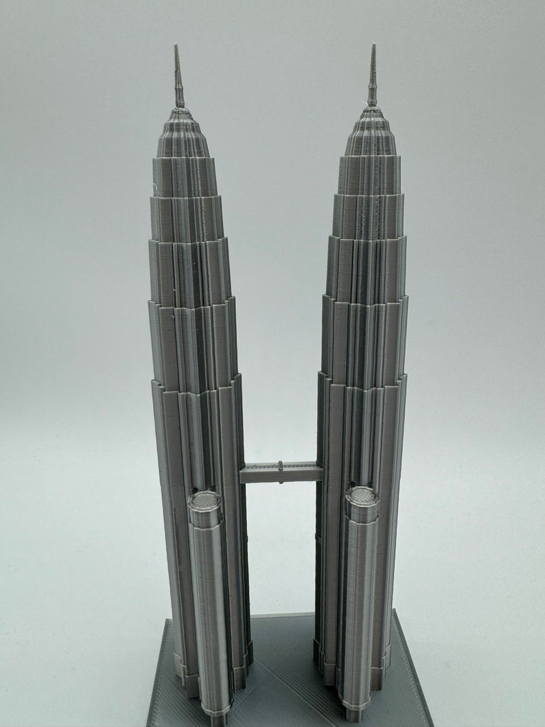 May include: A 3D printed model of the Petronas Towers in Kuala Lumpur, Malaysia. The model is made of gray plastic and features a bridge connecting the two towers.