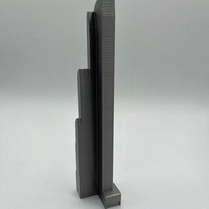 May include: A gray 3D-printed architectural model of a skyscraper. The model has a rectangular base and multiple vertical sections of varying heights, with detailed textured surfaces. The overall design is modern and minimalist.