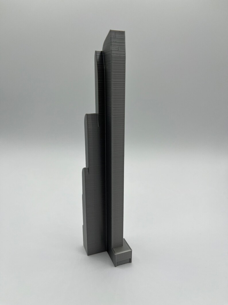 May include: A gray 3D-printed architectural model of a skyscraper. The model has a rectangular base and multiple vertical sections of varying heights, with detailed textured surfaces. The overall design is modern and minimalist.