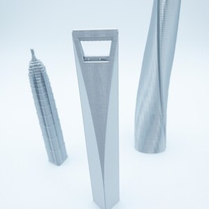 May include: Three silver 3D printed models of skyscrapers. The tallest model is a twisted skyscraper, the middle model is a tall, thin skyscraper with a hole in the middle, and the shortest model is a rectangular skyscraper.