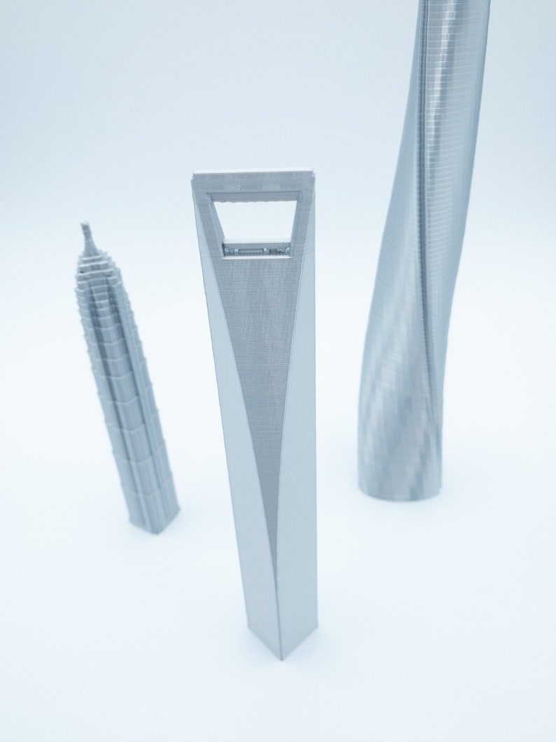 May include: Three silver 3D printed models of skyscrapers. The tallest model is a twisted skyscraper, the middle model is a tall, thin skyscraper with a hole in the middle, and the shortest model is a rectangular skyscraper.