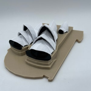May include: A white 3D printed model of the Sydney Opera House on a brown base.